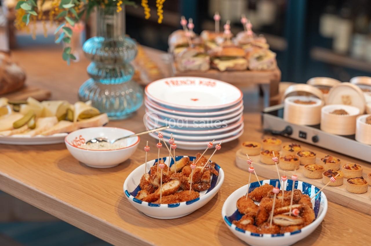 Assortiment de plats sur une table en bois, incluant bouchées apéritives, sandwichs, et vaisselle empilée.