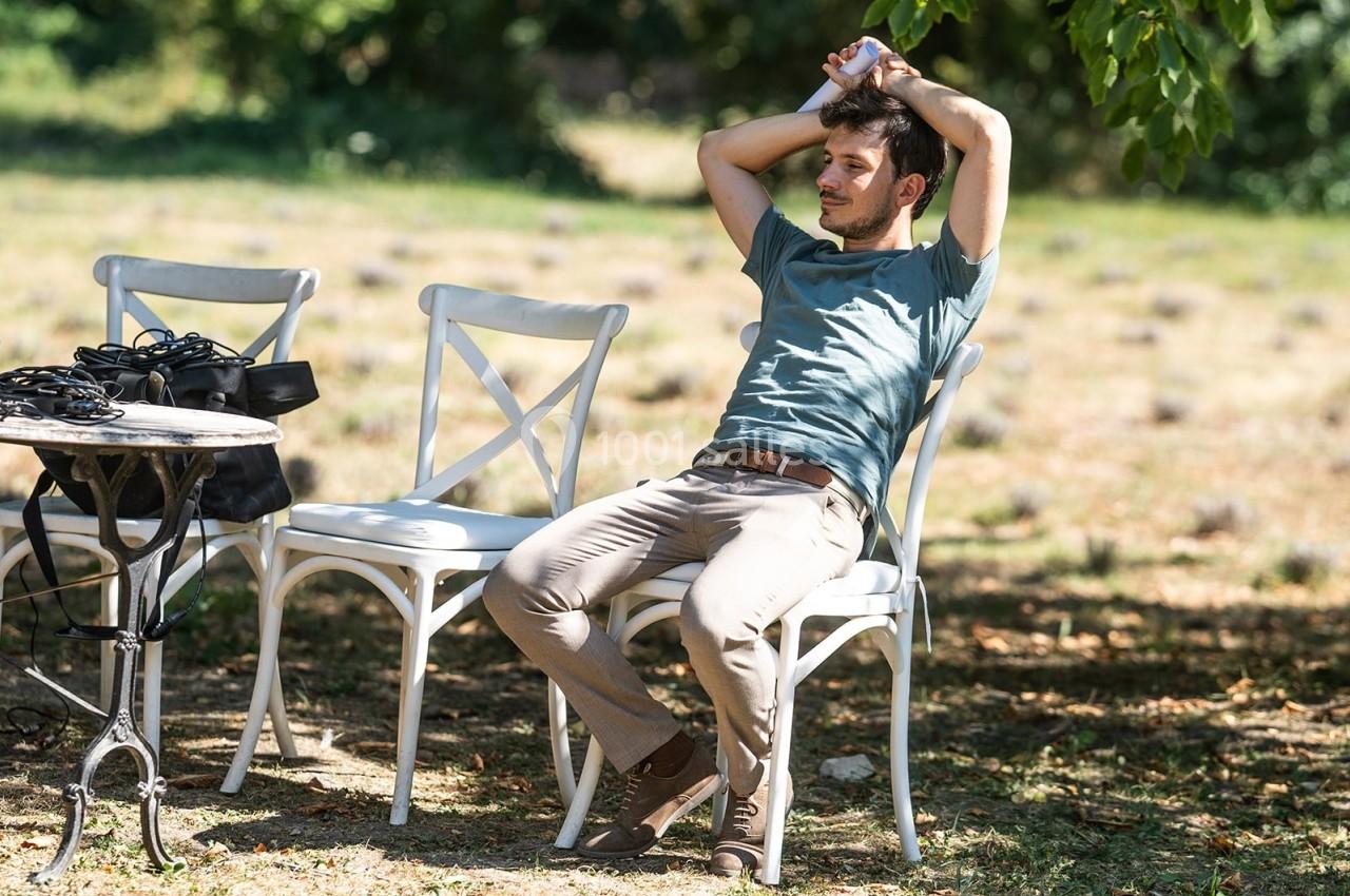 Un homme assis sur une chaise blanche dans un jardin ombragé, les bras derrière la tête, semblant se détendre.