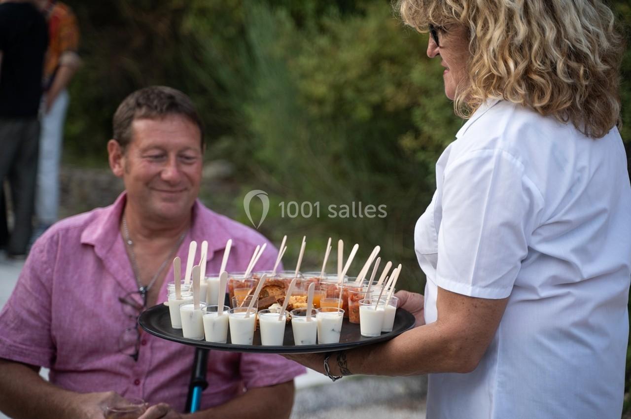 Une serveuse présente un plateau de verrines salées et sucrées à un homme assis en extérieur.