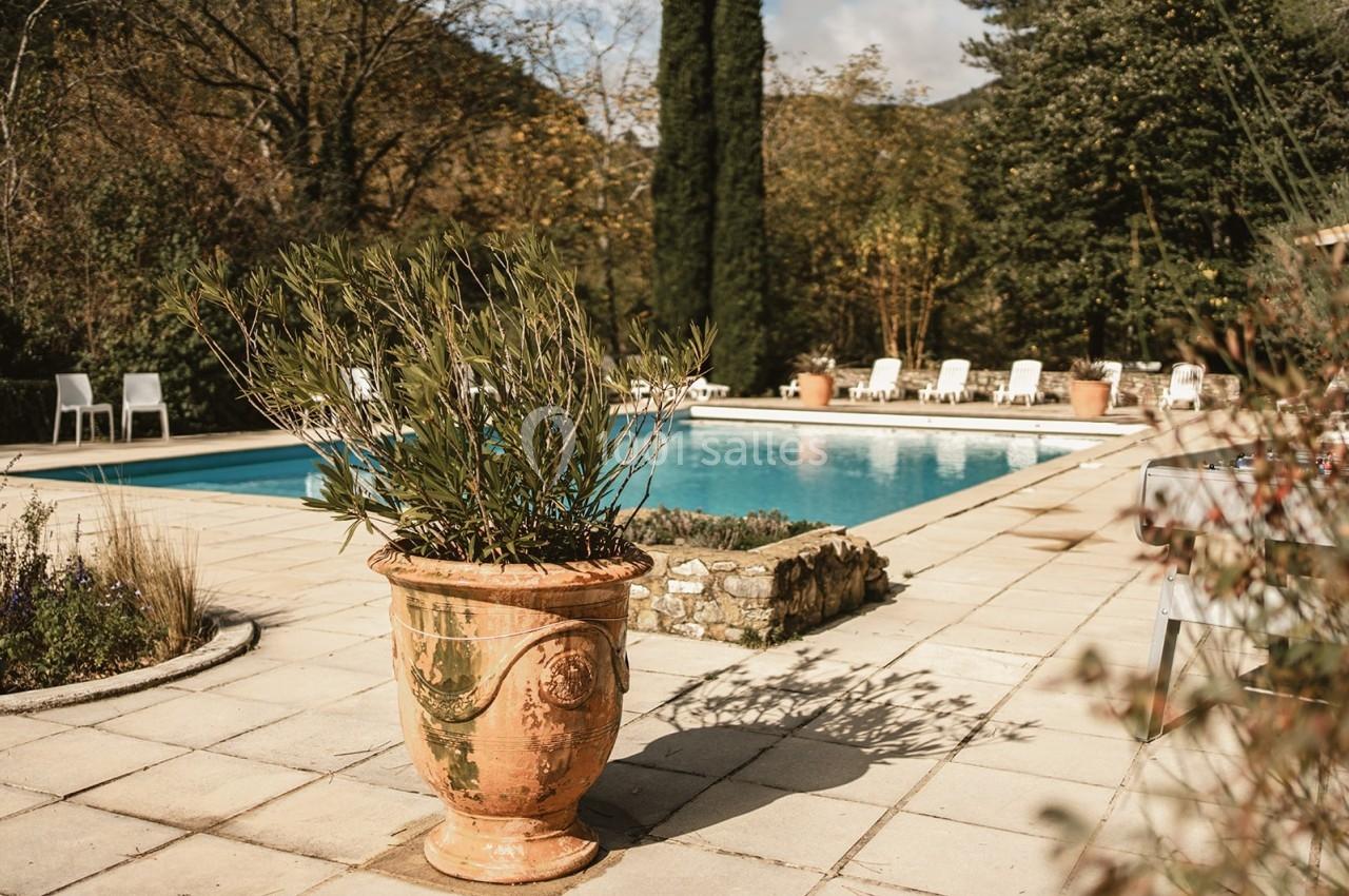 Piscine extérieure entourée de chaises longues, avec un grand pot en terre cuite et des arbres en arrière-plan.