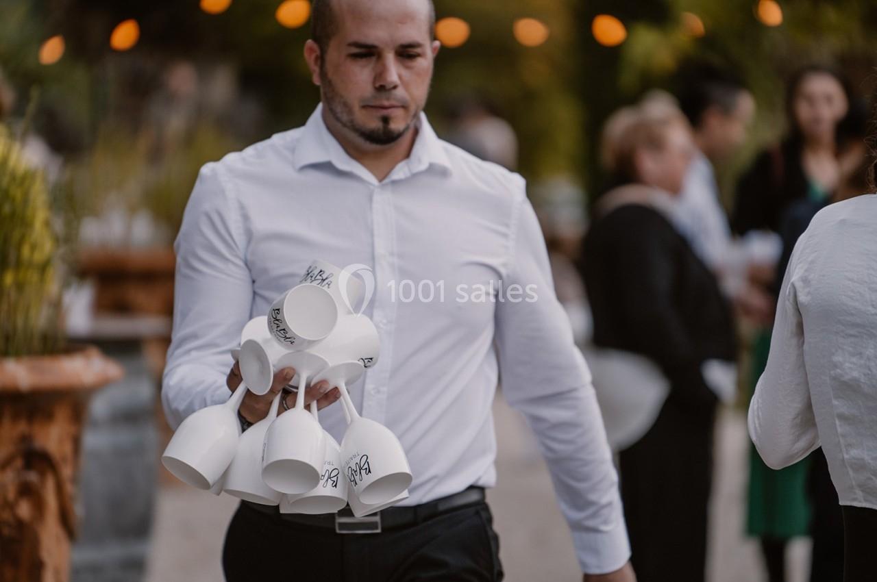 Un homme en chemise blanche porte plusieurs tasses blanches empilées par leurs anses lors d'un événement en extérieur.