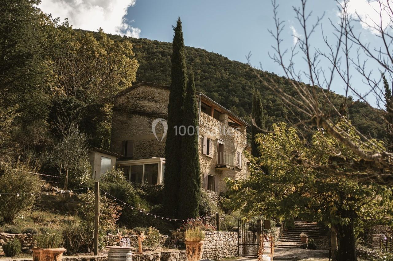 Maison en pierre entourée de cyprès et de végétation, située au pied d'une colline boisée sous un ciel partiellement nuageux.