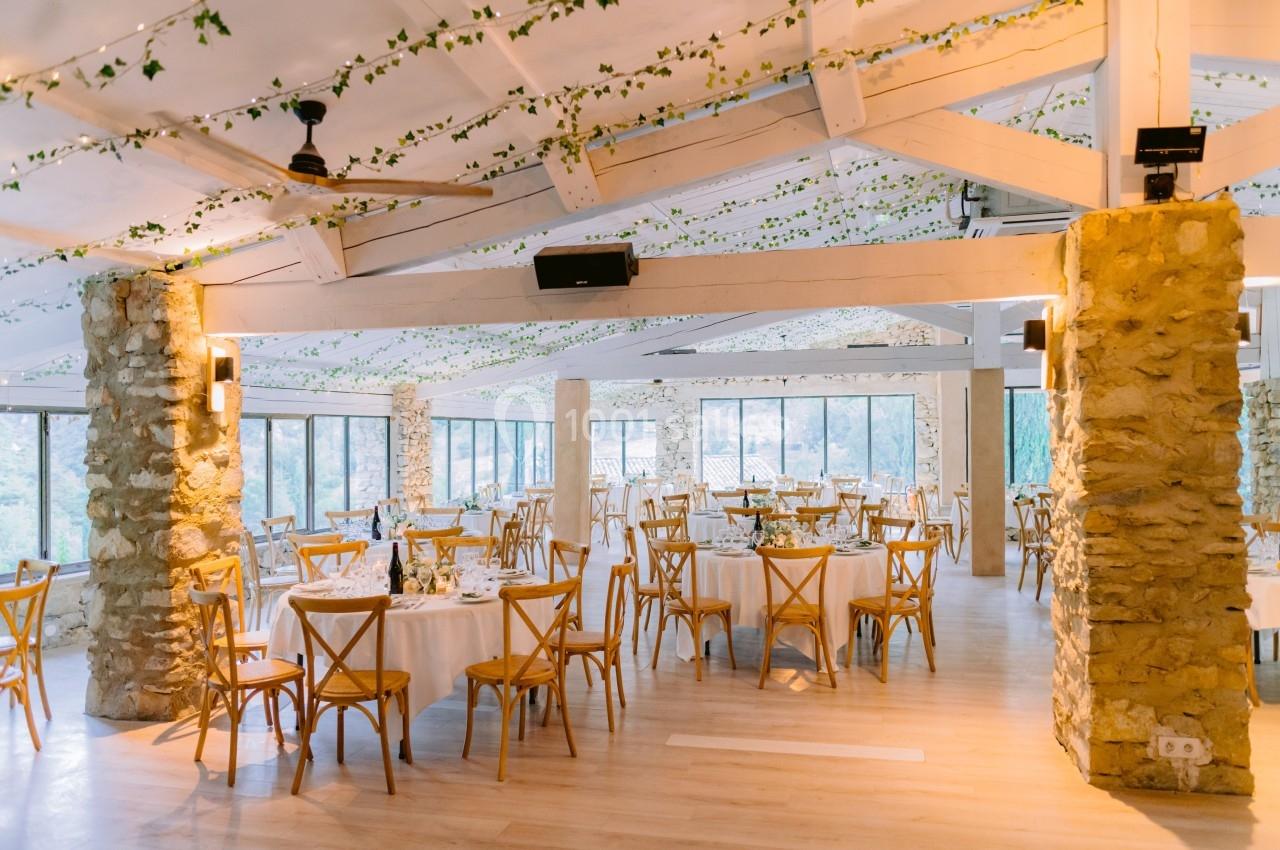 Salle de réception lumineuse avec des tables rondes dressées, des chaises en bois et une décoration végétale suspendue.
