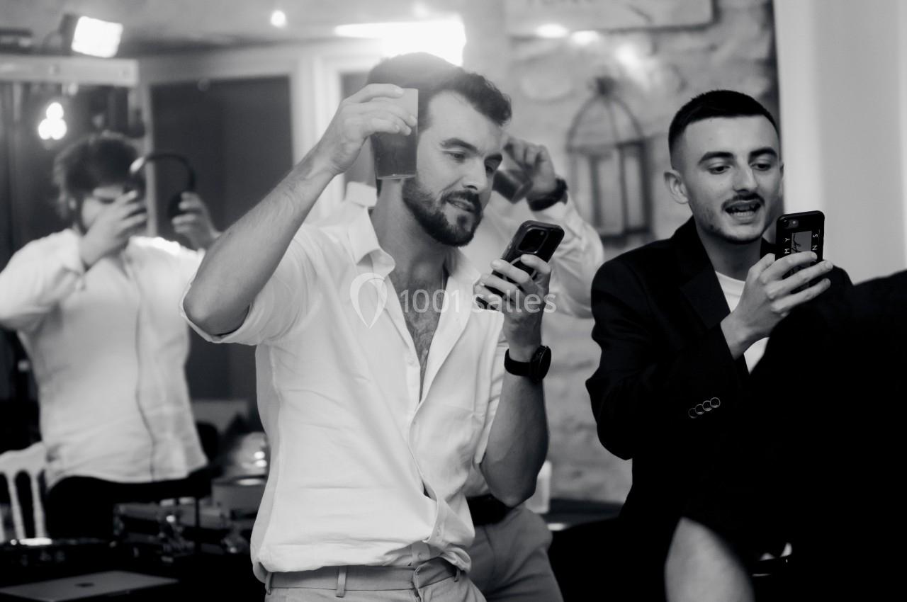 Des hommes debout dans une ambiance festive, utilisant leurs téléphones, avec un fond flou de décor intérieur.