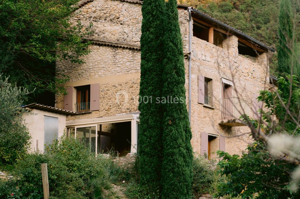 Groupe de personnes rassemblées devant une maison en pierre entourée de végétation, avec un cyprès au premier plan.