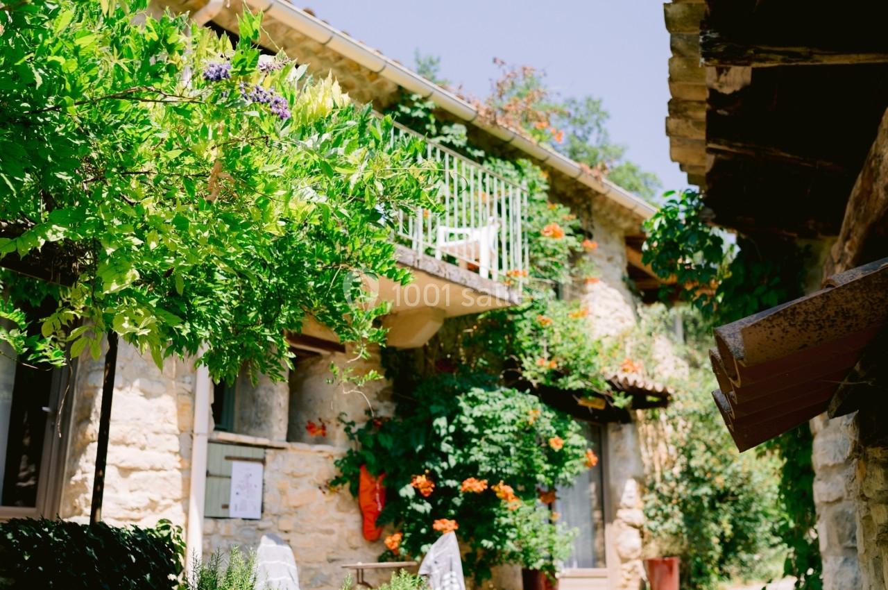 Façade en pierre d'une maison entourée de végétation, avec un balcon fleuri et un chemin en pierre.