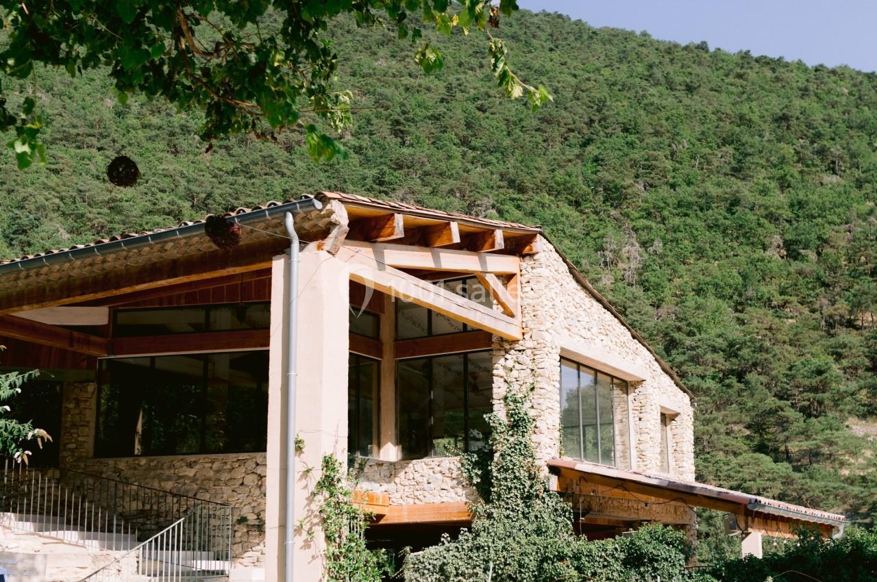 Bâtiment en pierre avec de grandes baies vitrées, entouré de verdure et situé au pied d'une colline boisée.