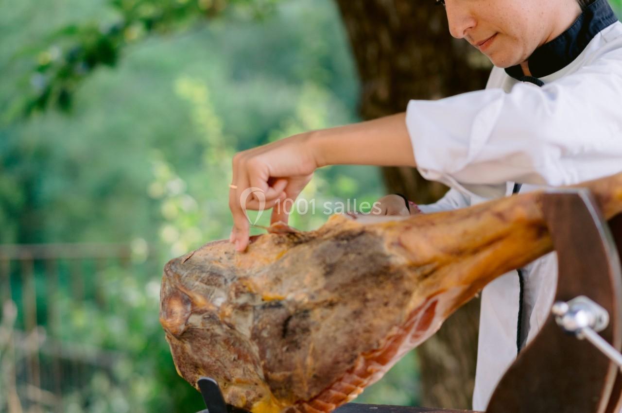 Une femme en tenue de chef découpe un jambon cru sur un support en bois, en extérieur sous un arbre.