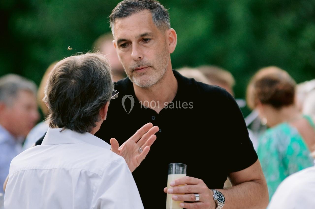 Deux hommes discutent debout lors d'un événement en plein air, l'un tenant un verre à la main.