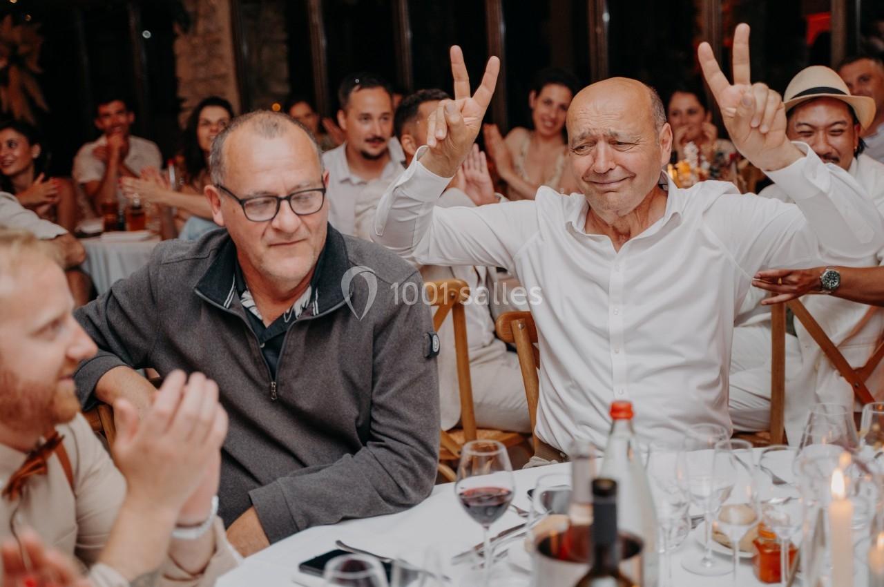 Deux hommes assis à une table lors d'un dîner festif, l'un souriant et levant les bras en signe de victoire.