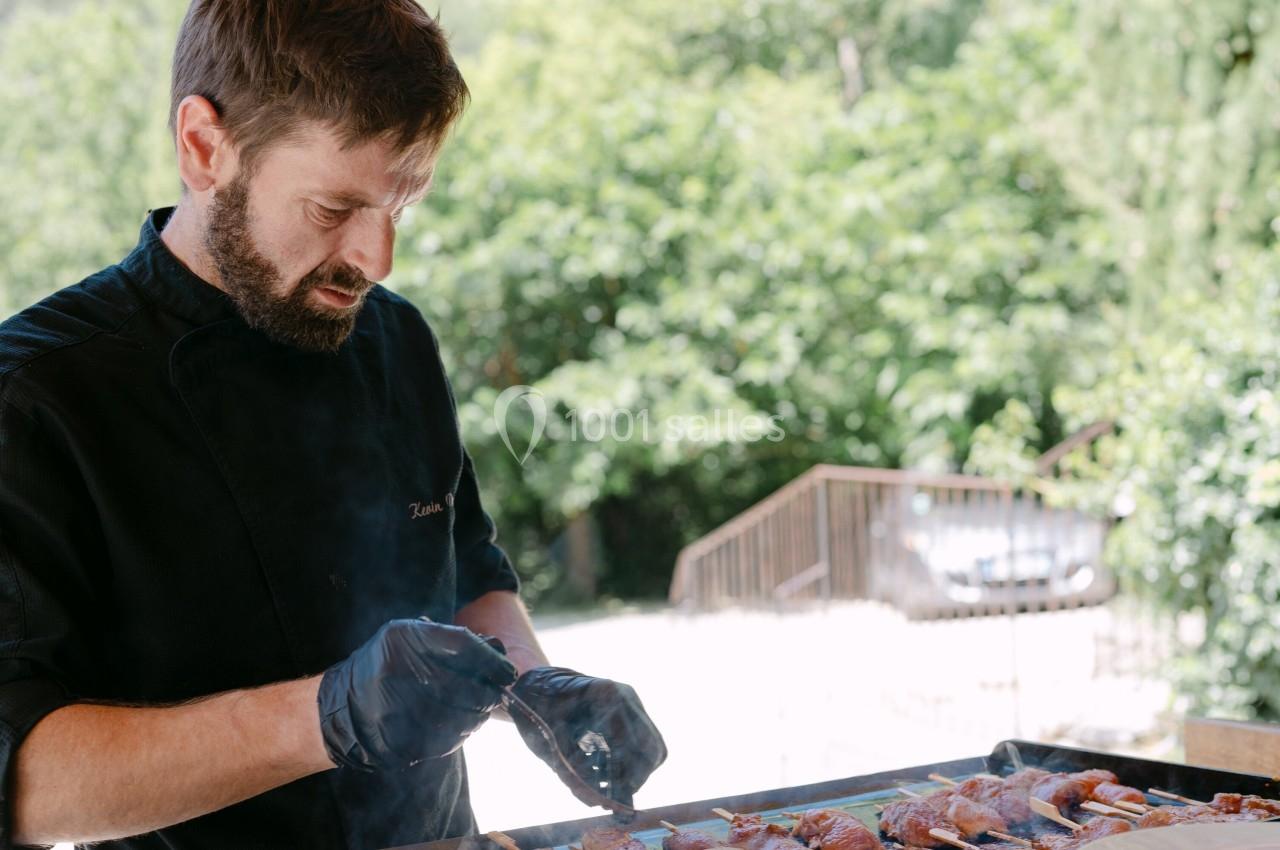 Un cuisinier prépare des brochettes sur un gril en extérieur, avec des assiettes de charcuterie au premier plan.