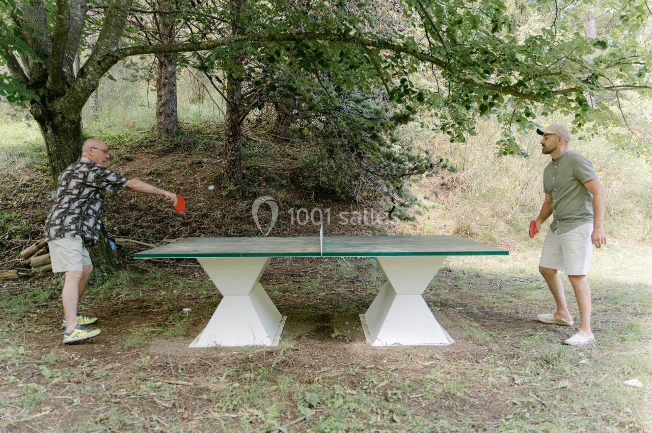 Deux hommes jouent au ping-pong en plein air sur une table située dans un cadre naturel boisé.