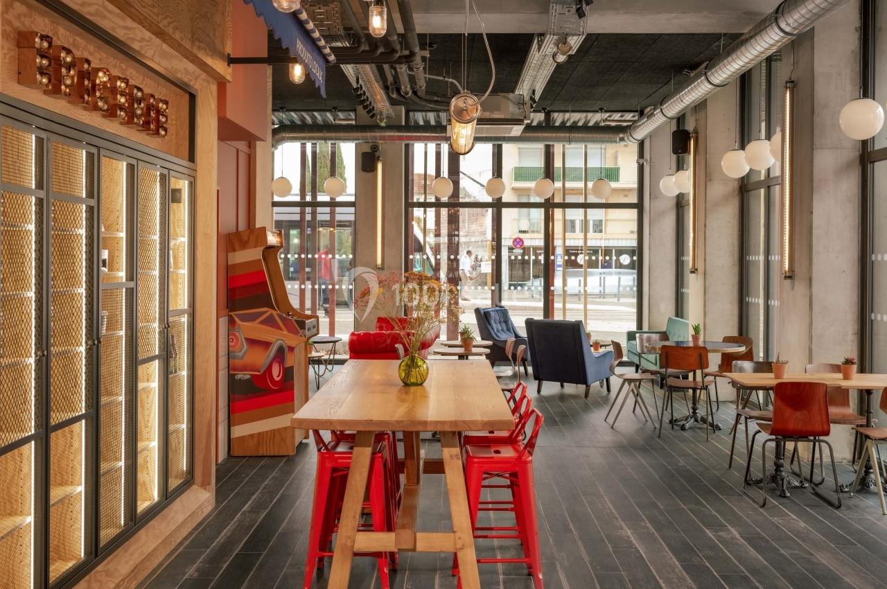 Salle lumineuse avec tables en bois, chaises colorées, grandes fenêtres et décoration moderne industrielle.