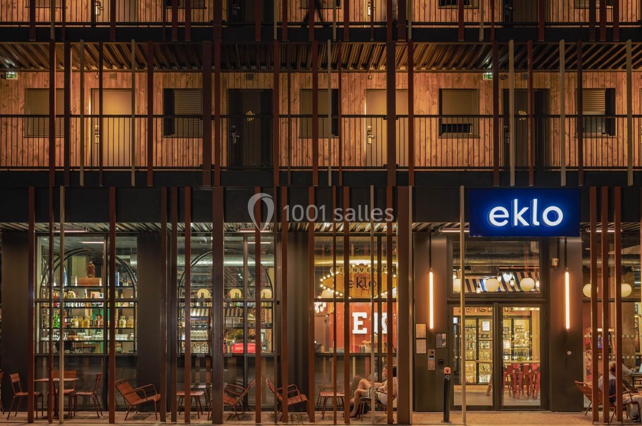 Façade d'un bâtiment moderne avec une enseigne lumineuse ’eklo’, des baies vitrées et des tables en terrasse.