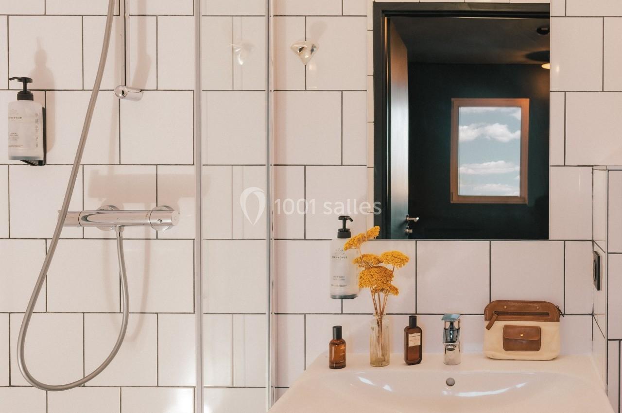 Salle de bain avec carrelage blanc, douche, lavabo, miroir, flacons et fleurs jaunes en décoration.