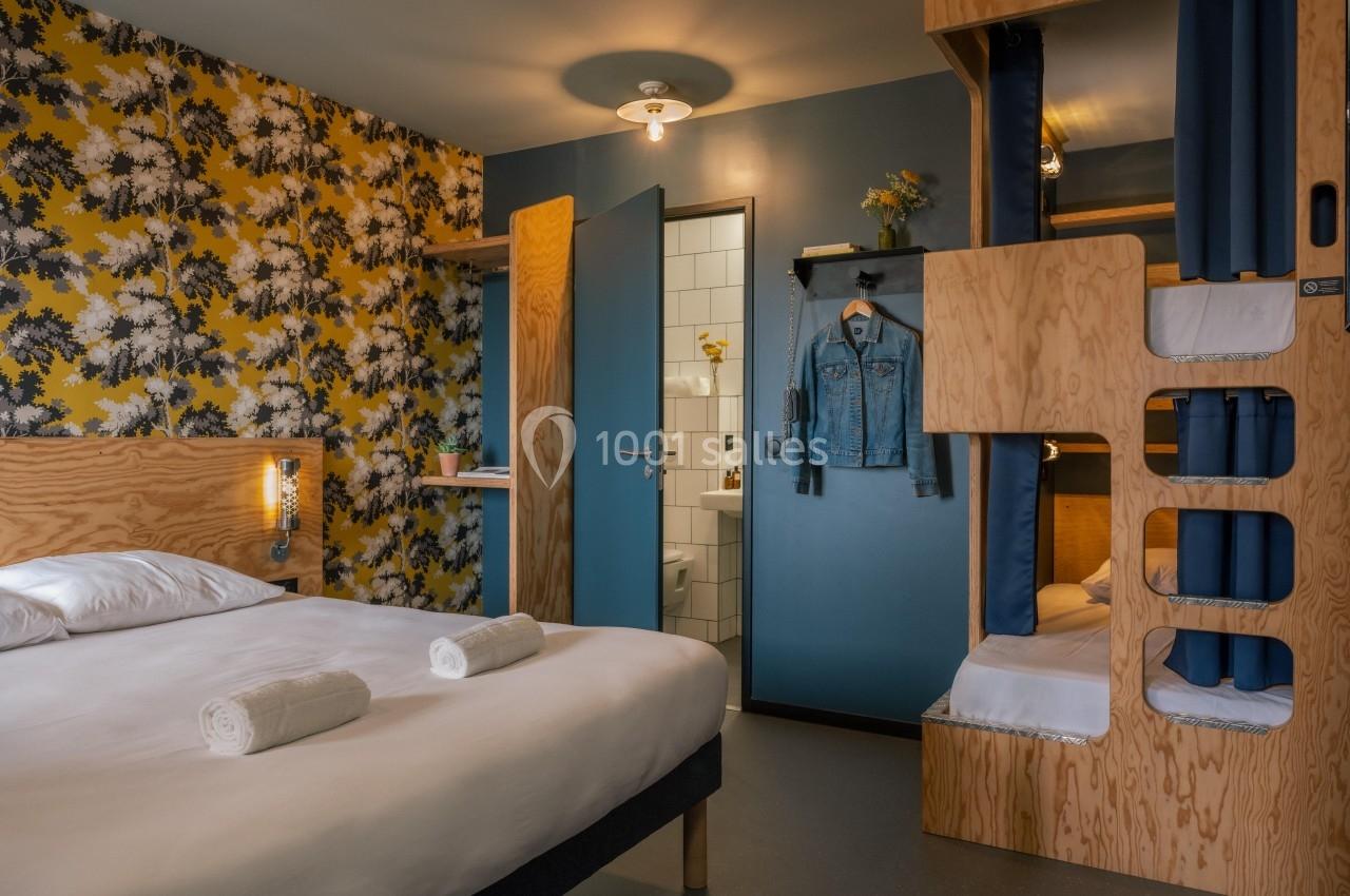Chambre d'hôtel avec lit double, lits superposés en bois, mur fleuri, et salle de bain visible à travers une porte ouverte.
