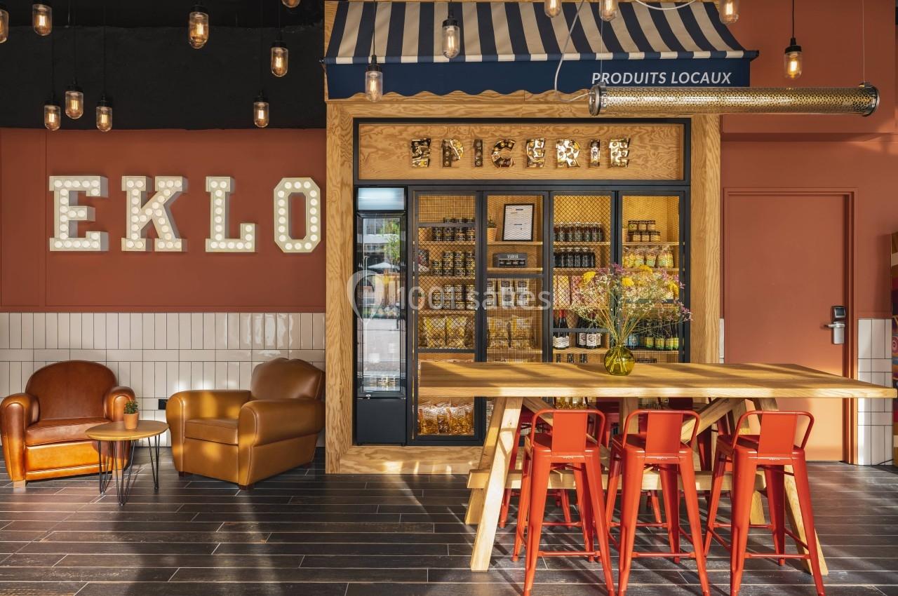 Coin boutique avec étagères de produits locaux, table en bois, chaises rouges et fauteuils dans un décor chaleureux.
