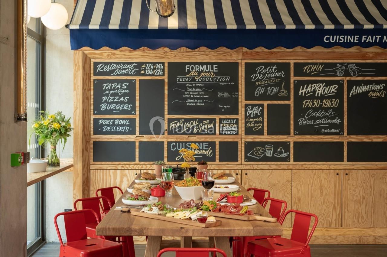 Salle de restaurant avec table en bois dressée pour un repas, ardoises murales affichant menus et offres, chaises rouges.