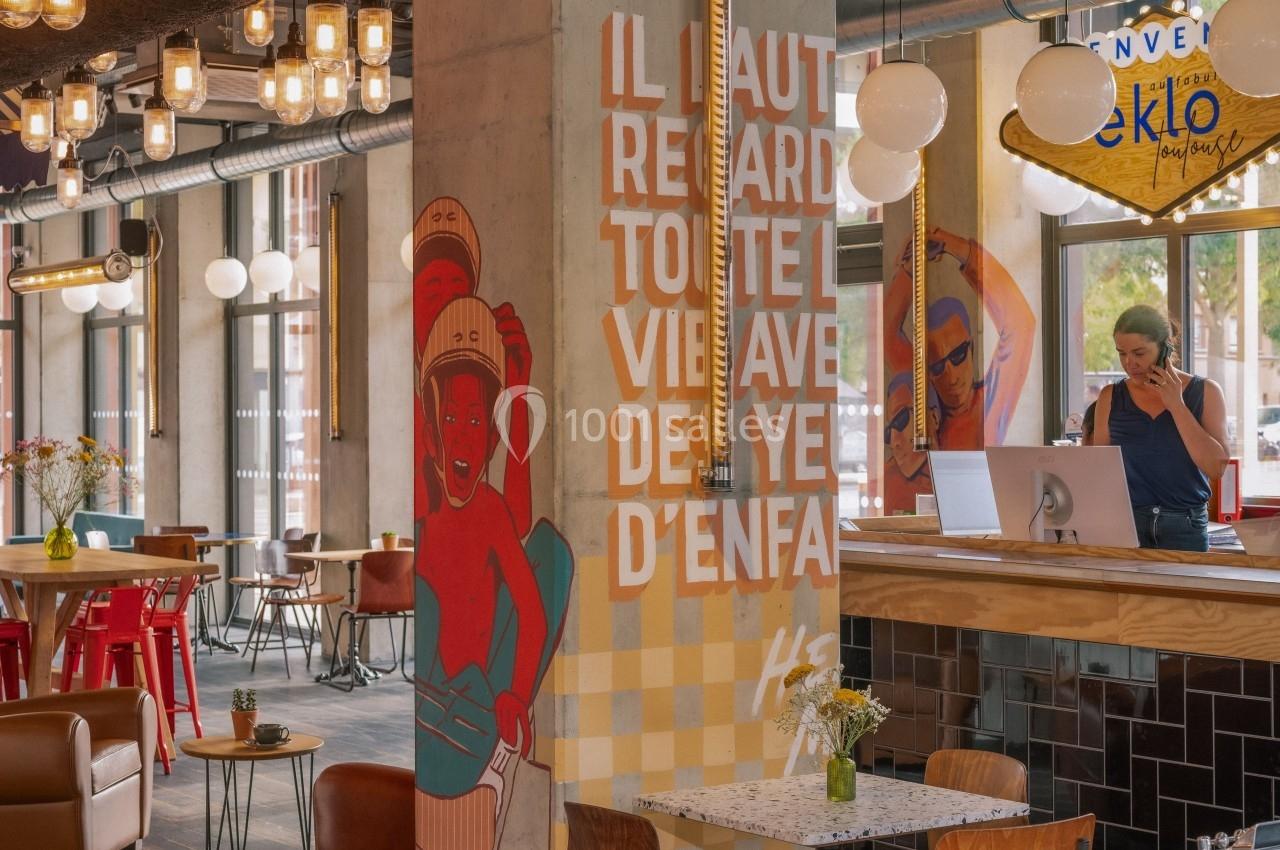 Intérieur d'un café moderne avec une fresque colorée, un comptoir en bois et une personne au téléphone derrière le comptoir.