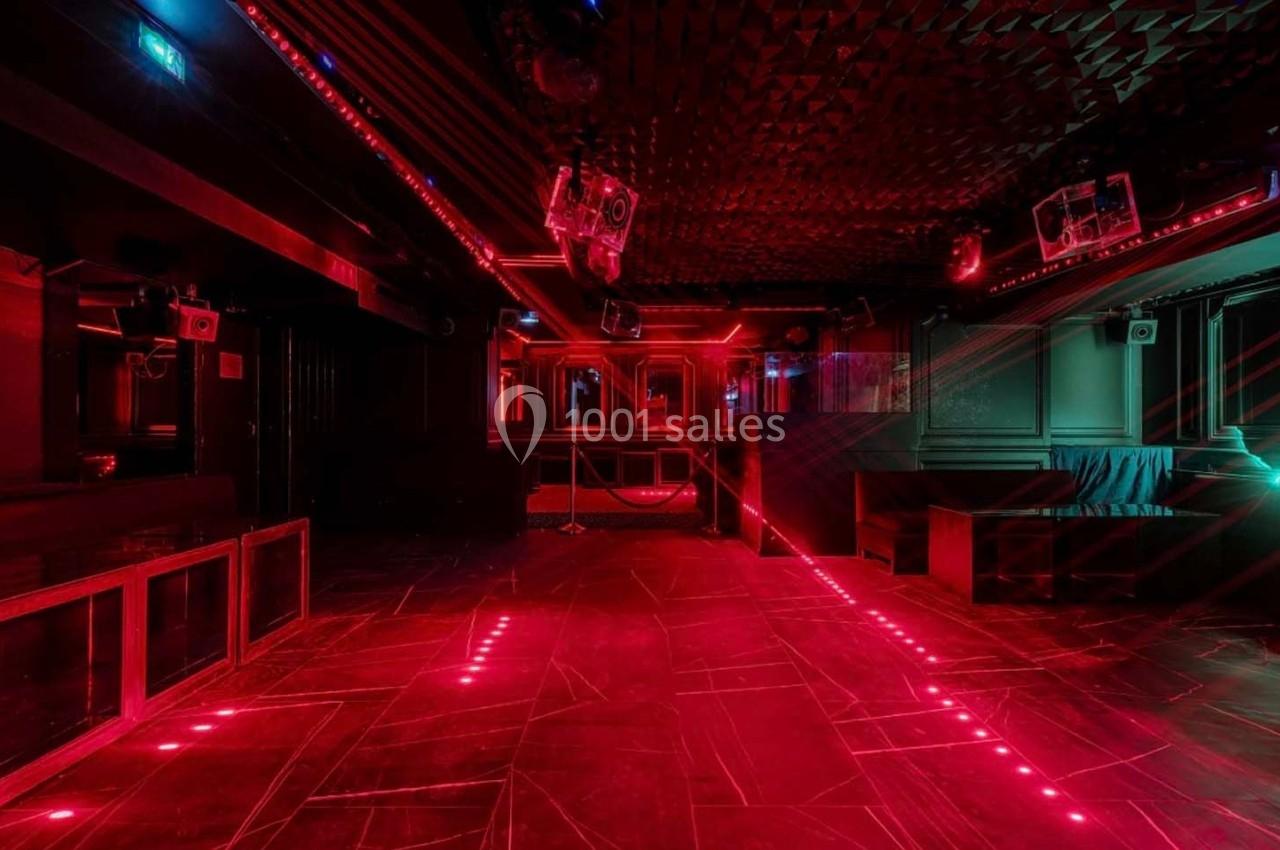 Salle de club vide avec éclairage rouge, murs sombres, sol en motifs géométriques et équipements audio visibles.