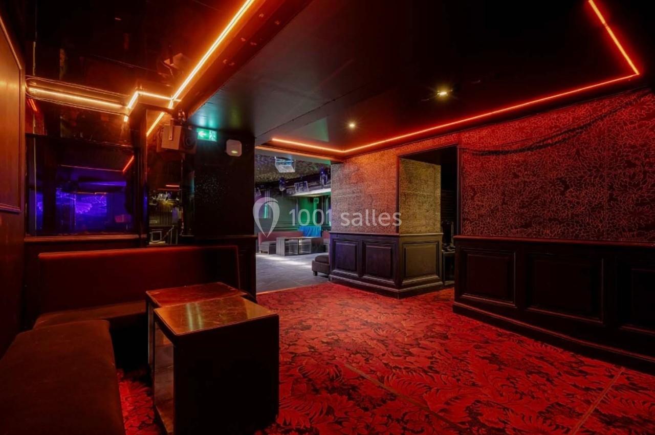 Intérieur d'un club avec éclairage rouge et violet, banquettes, tables basses et moquette à motifs.