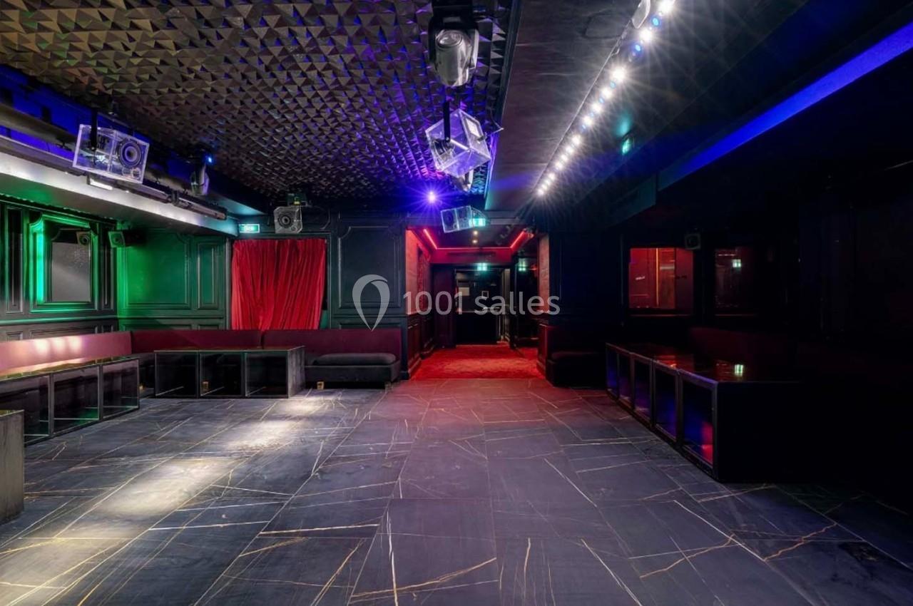 Salle sombre avec éclairage coloré, murs noirs, rideau rouge, banquettes et sol en carrelage sombre.