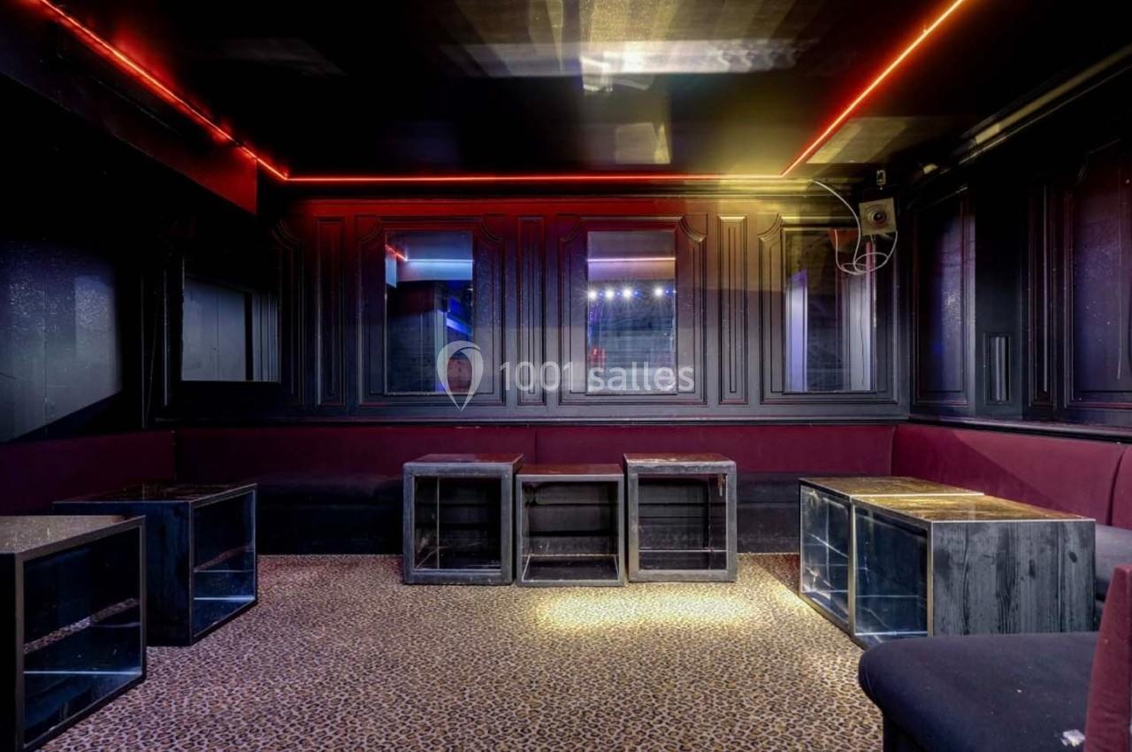 Salle sombre avec banquettes rouges, tables basses en bois et éclairage LED rouge et jaune au plafond.