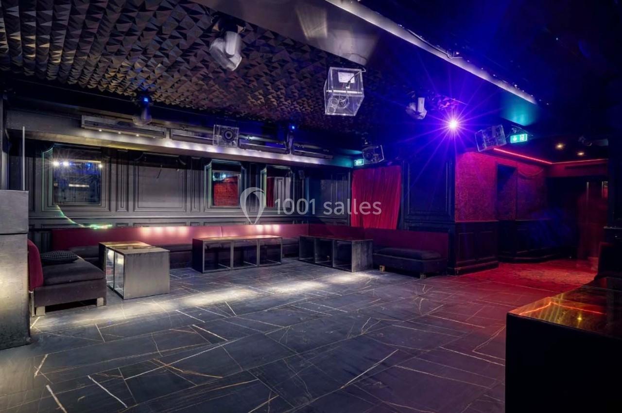 Salle de club vide avec éclairage tamisé, murs sombres, banquettes et tables basses disposées autour de la pièce.