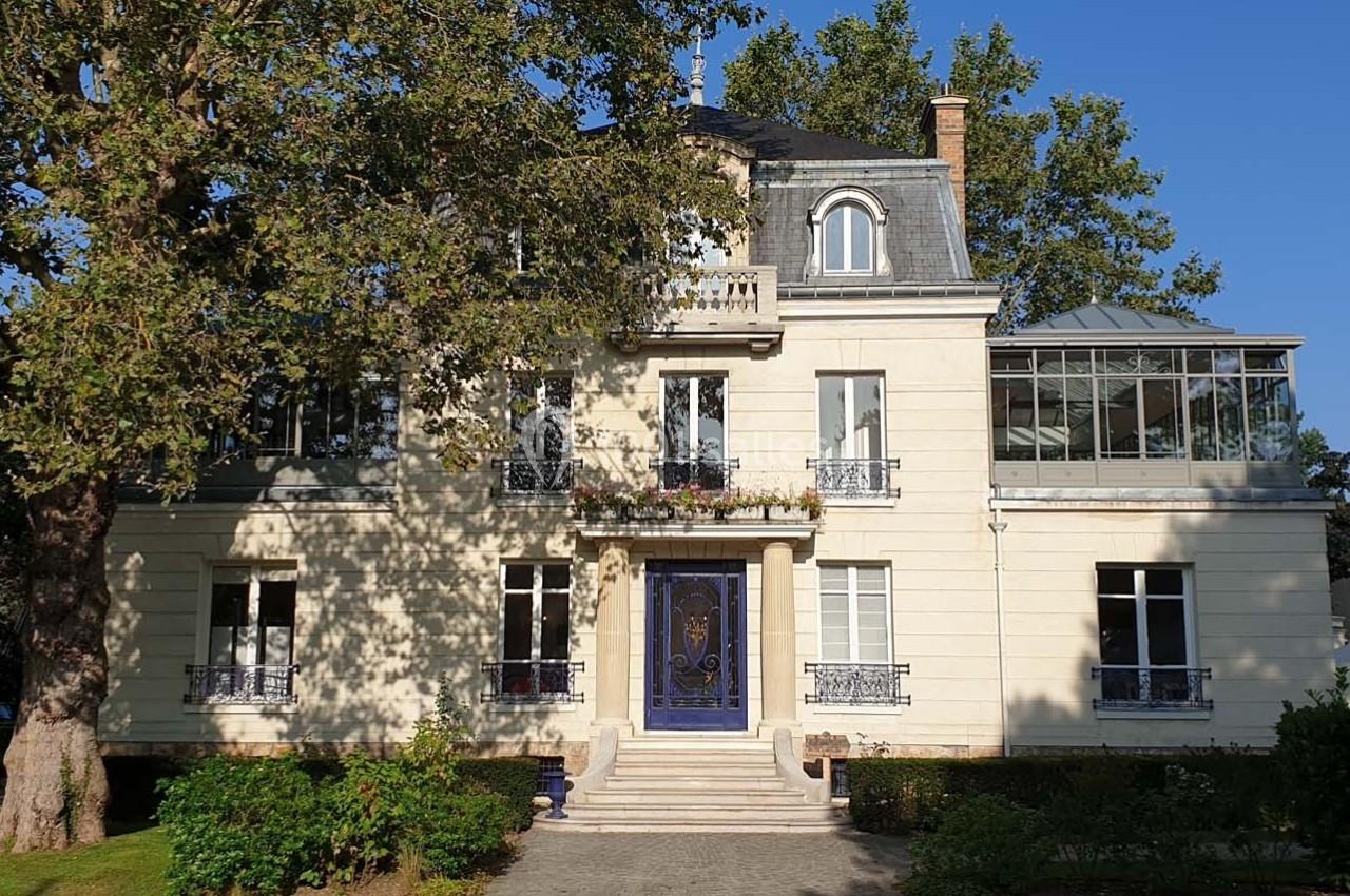 Façade d'une grande maison bourgeoise avec un toit mansardé, entourée d'arbres et d'un jardin.