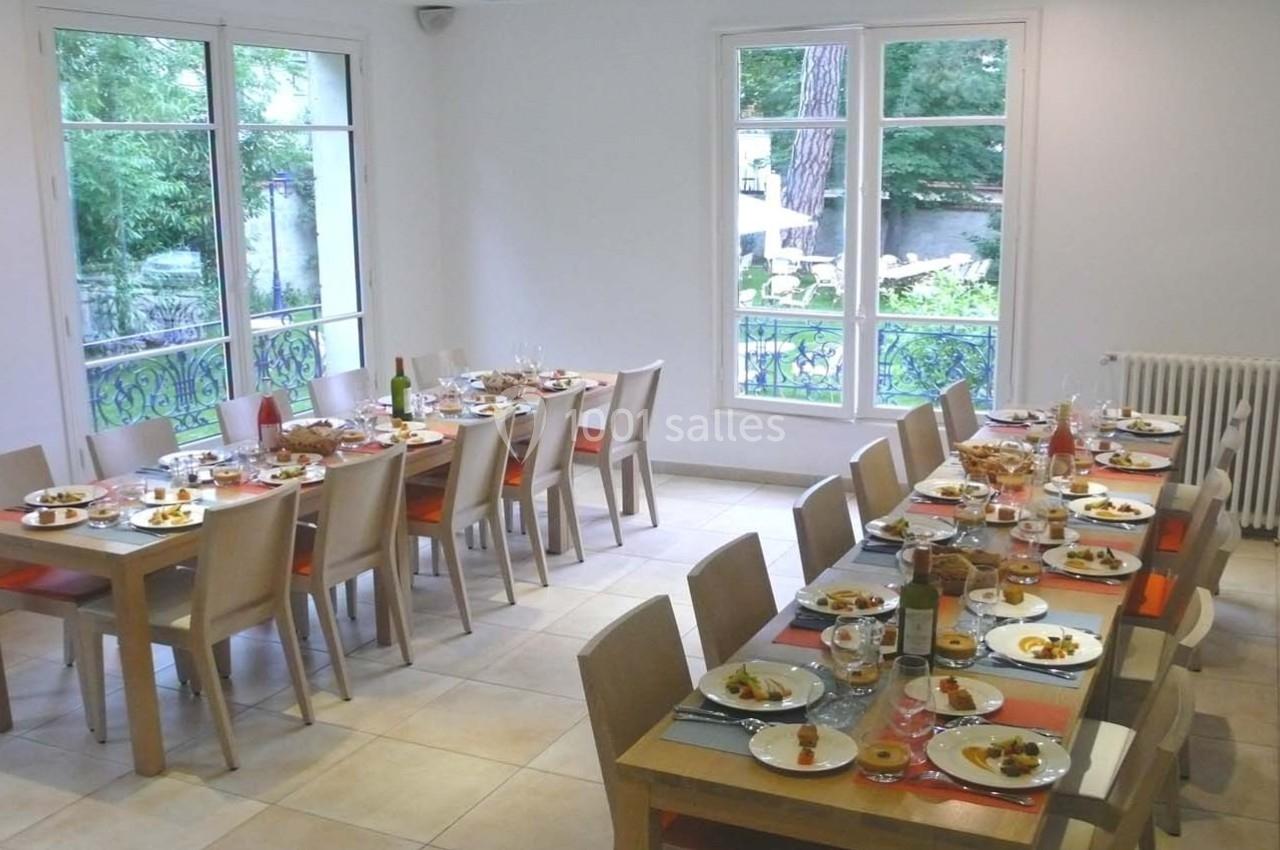 Salle à manger lumineuse avec deux grandes tables dressées pour un repas, entourées de chaises en bois clair.