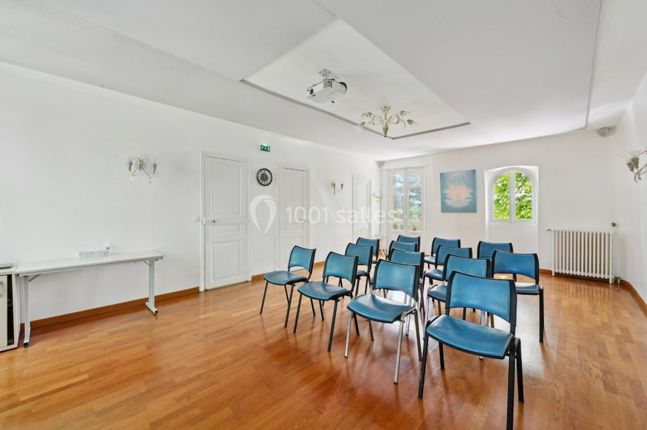 Salle lumineuse avec parquet, chaises bleues disposées en rangées, table blanche et fenêtres donnant sur un espace vert.