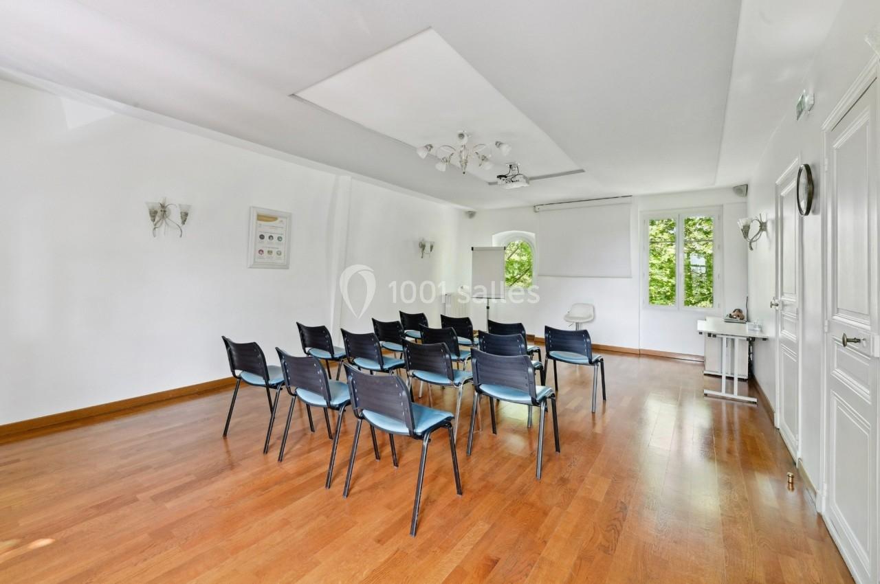 Salle lumineuse avec parquet, rangée de chaises noires, tableau blanc et fenêtres donnant sur un espace vert.