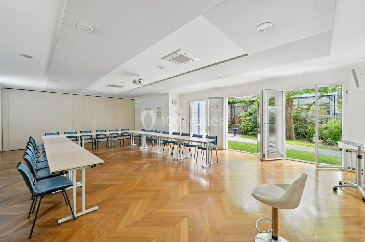Salle lumineuse avec parquet, tables et chaises disposées en U, grandes baies vitrées ouvertes sur un jardin.