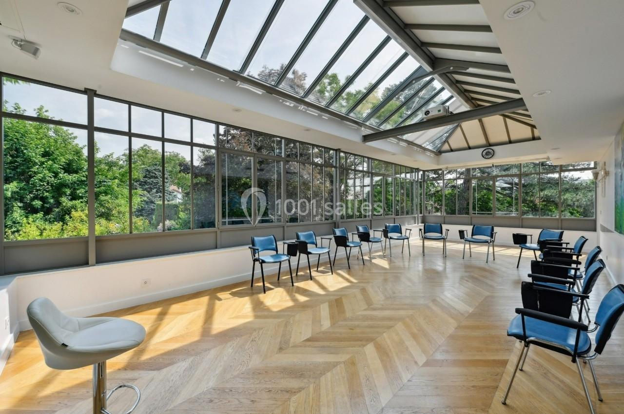 Salle lumineuse avec parquet en bois, grandes baies vitrées, chaises disposées en cercle et vue sur un jardin verdoyant.