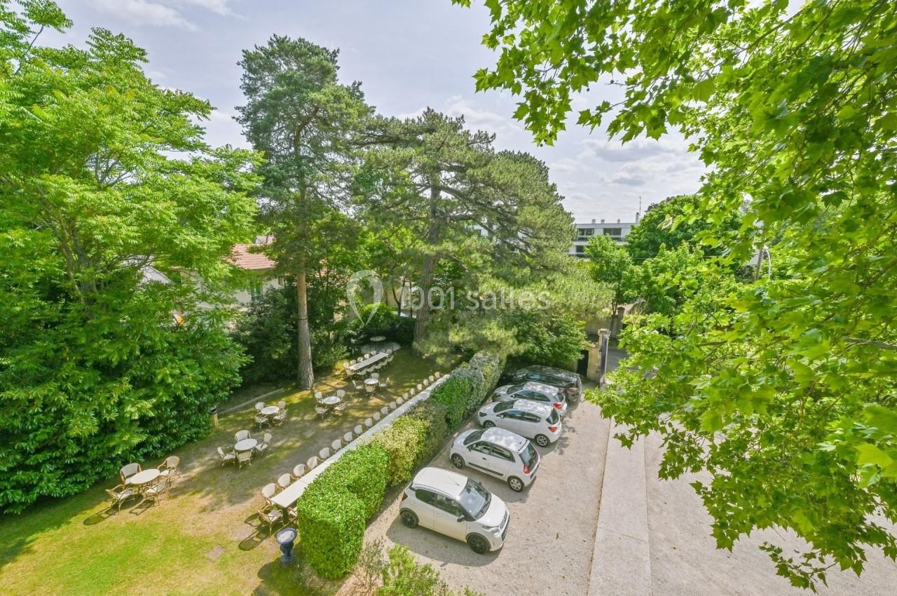 Vue d'un jardin arboré avec des tables et chaises, jouxtant un parking contenant plusieurs voitures.