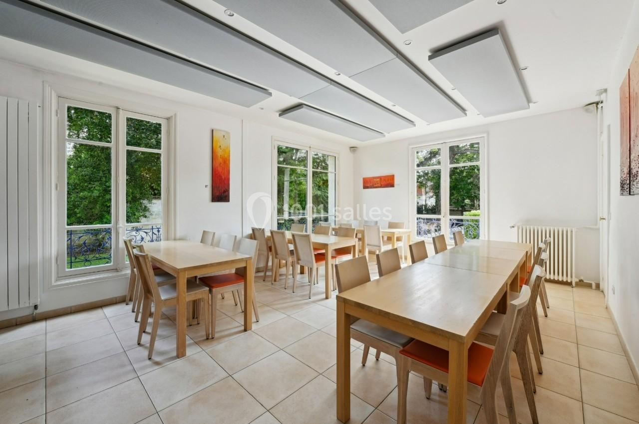 Salle lumineuse avec grandes fenêtres, tables en bois clair, chaises assorties et vue sur un jardin verdoyant.