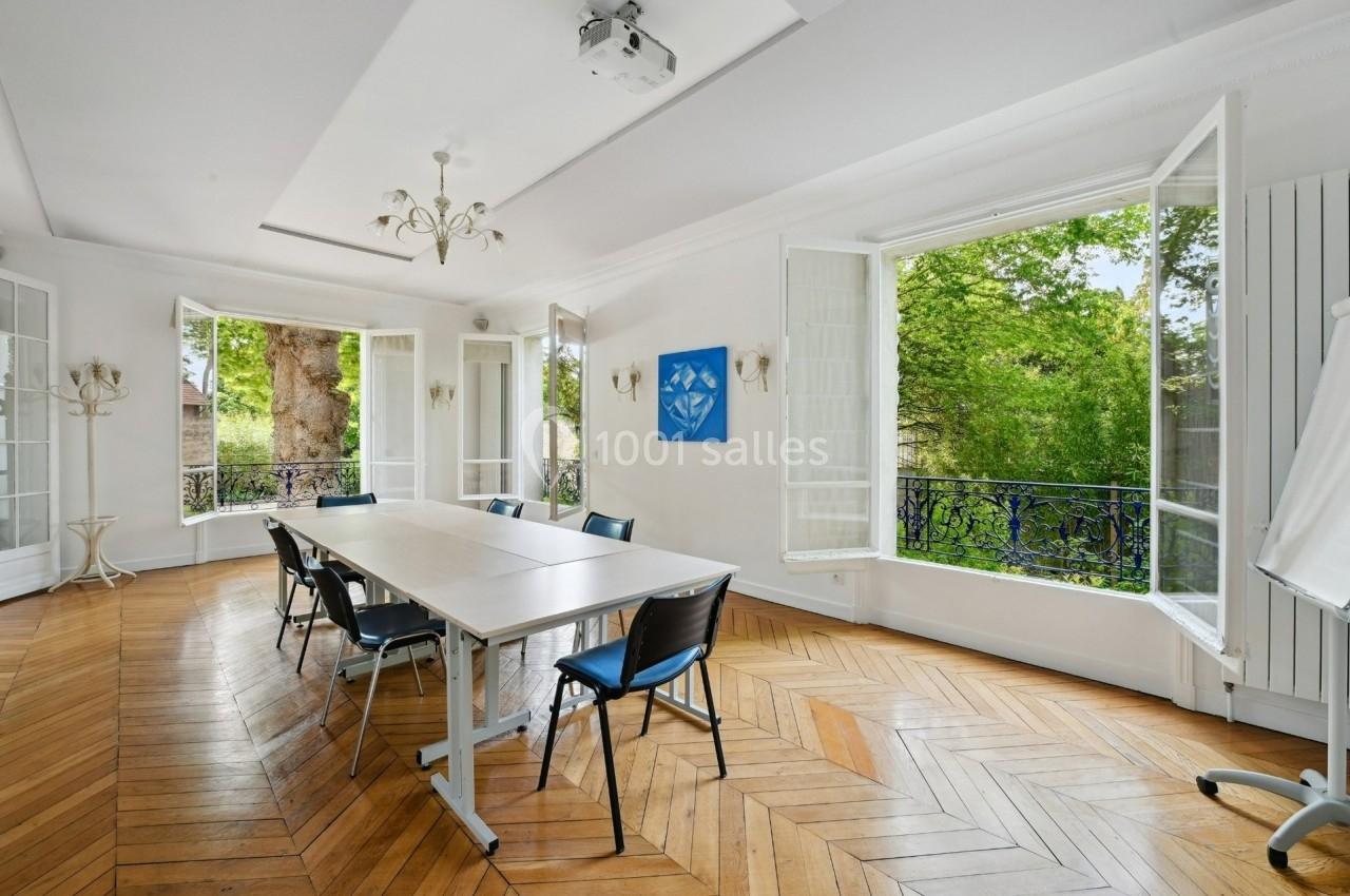 Salle de réunion lumineuse avec parquet en bois, table rectangulaire, chaises noires et grandes fenêtres ouvertes sur un…