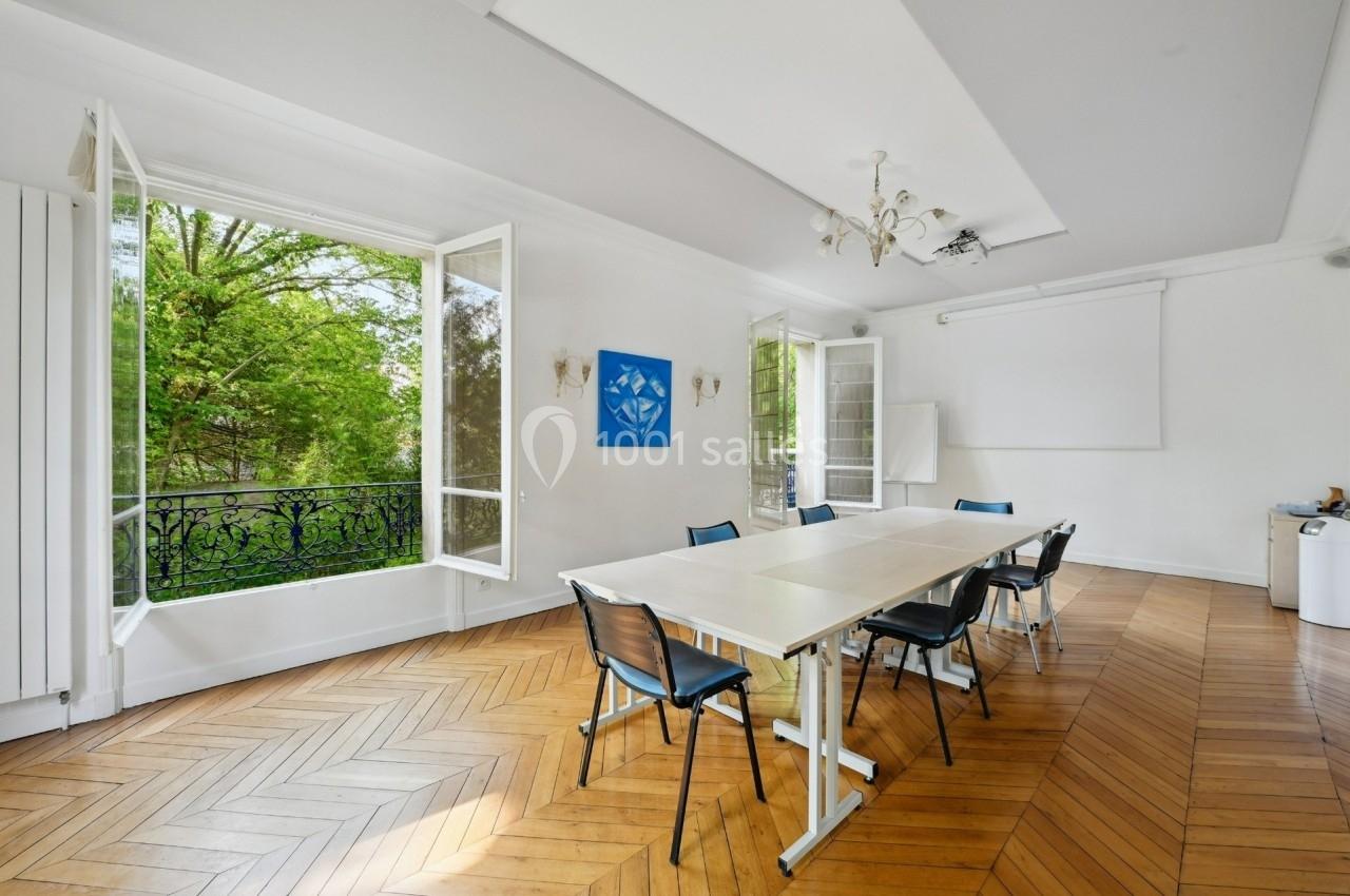 Salle de réunion lumineuse avec table, chaises, parquet en bois, fenêtres ouvertes sur un jardin verdoyant.