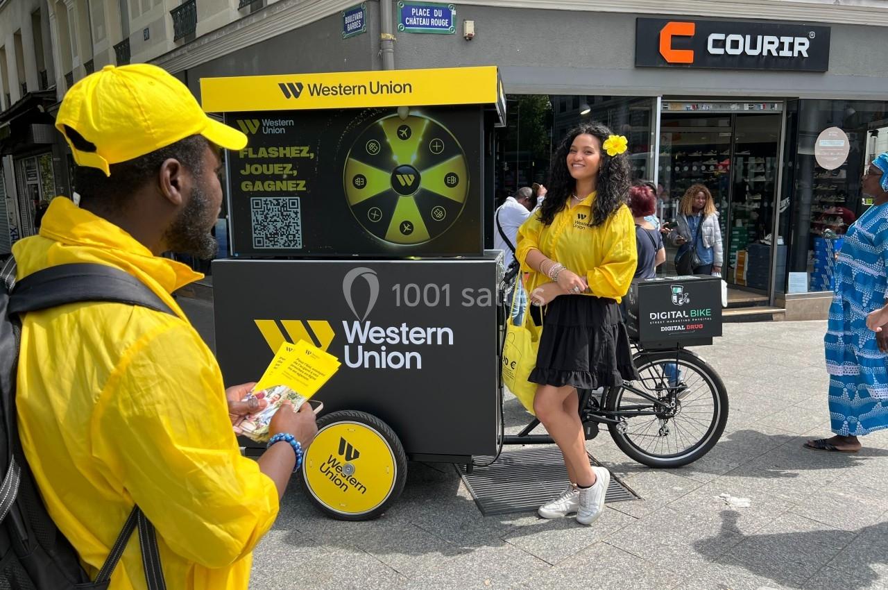 Deux personnes en tenue jaune près d'un vélo promotionnel Western Union dans une rue commerçante.