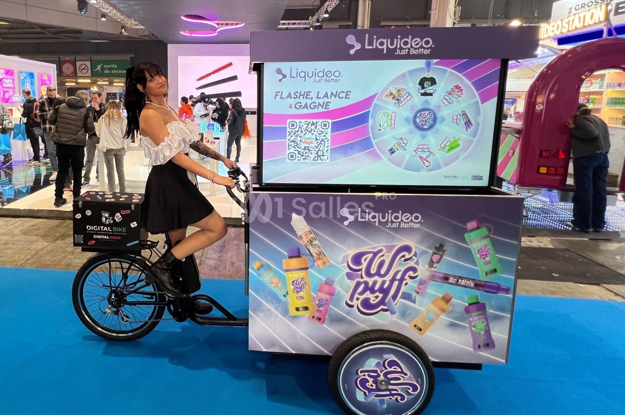 Femme posant à côté d'un vélo publicitaire présentant des produits de vapotage dans un salon ou un événement.