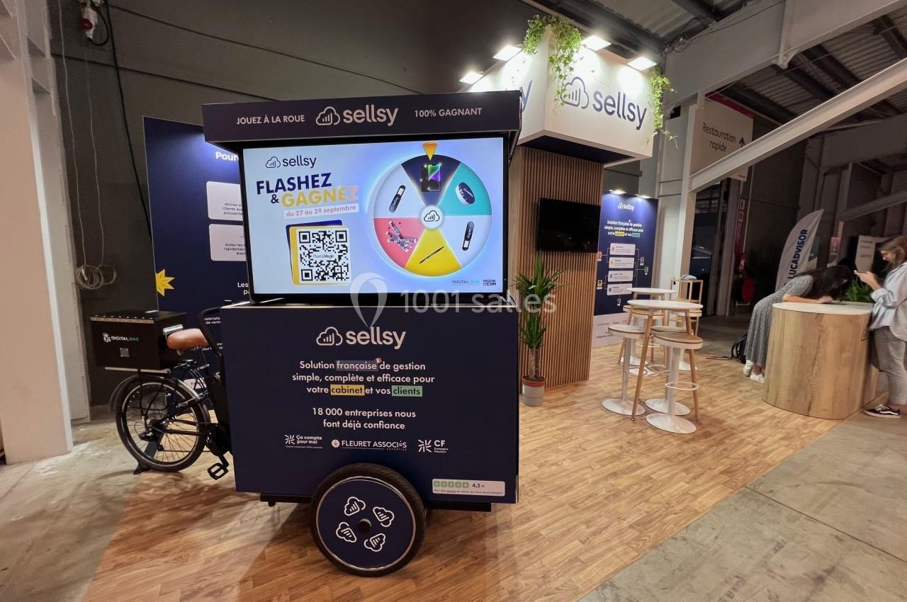Stand d'exposition avec un écran interactif, une roue de loterie et un triporteur, dans un espace intérieur éclairé.