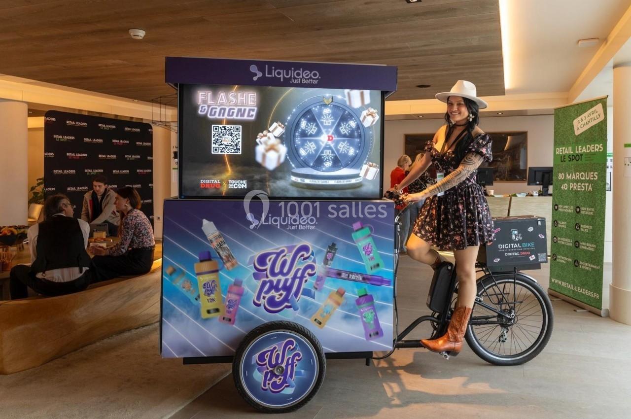 Une femme en tenue western pose près d'un triporteur promotionnel de produits de vape dans un espace intérieur moderne.