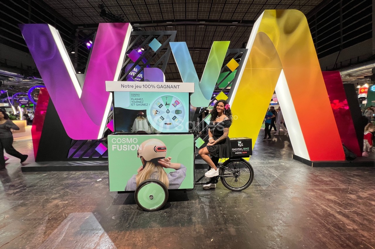Une femme pose près d'un triporteur promotionnel devant une structure colorée dans un espace événementiel.