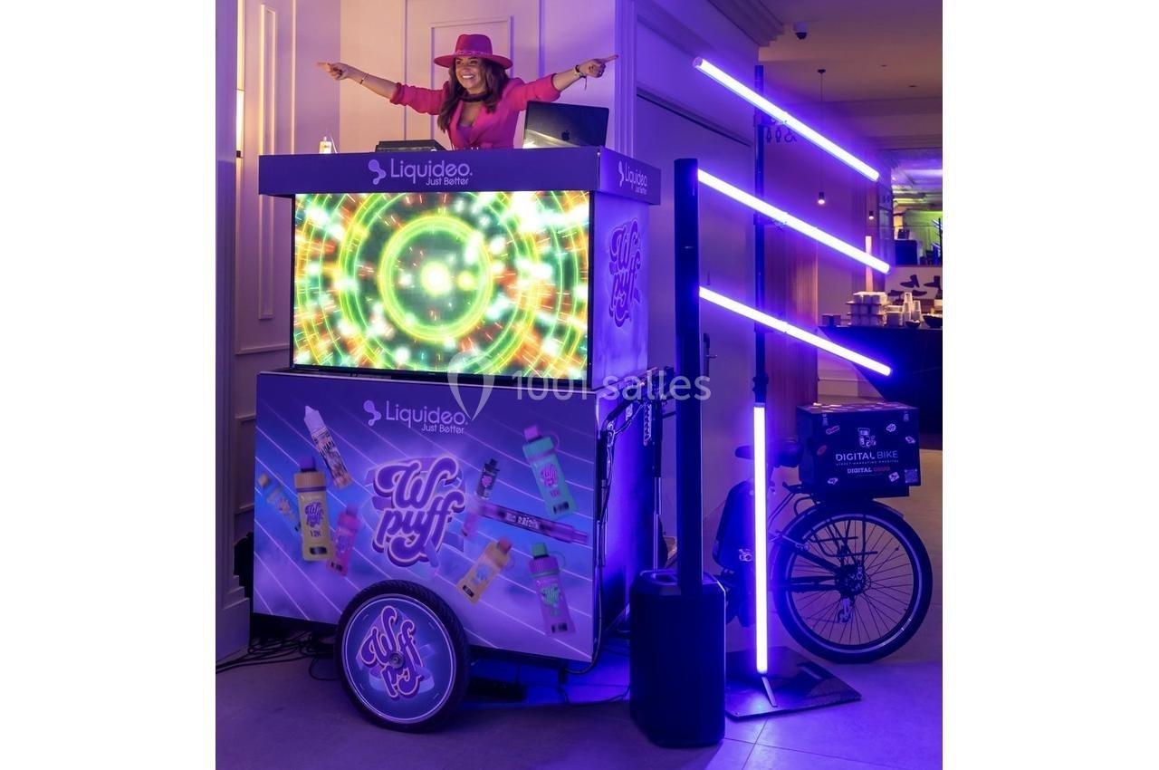 Femme souriante sur un stand lumineux décoré de motifs colorés, avec des tubes LED violets et un vélo intégré.
