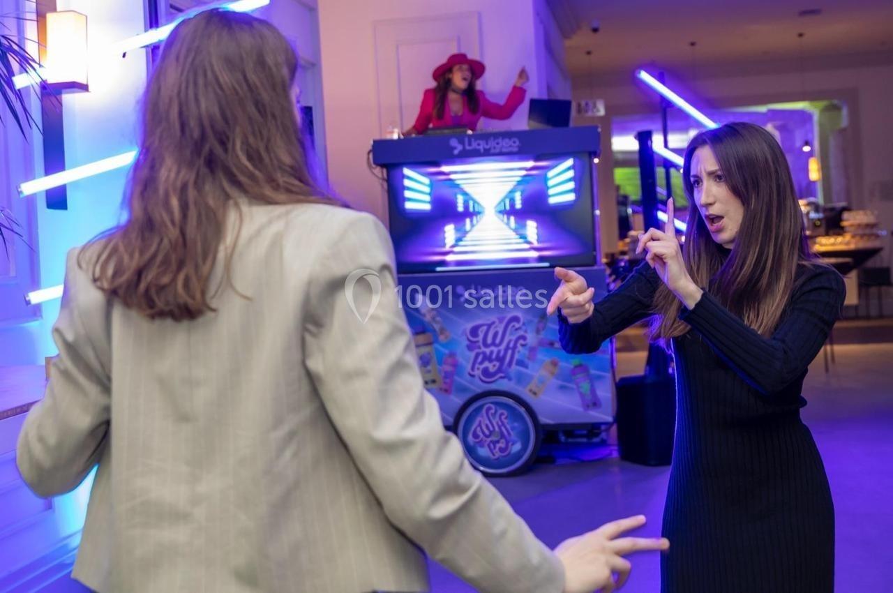 Deux personnes dansent dans un espace éclairé par des néons, avec une DJ en arrière-plan.