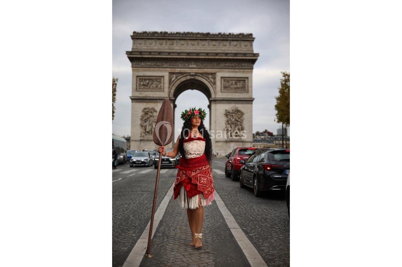 Femme en tenue traditionnelle polynésienne tenant une pagaie, posant devant l'Arc de Triomphe à Paris.