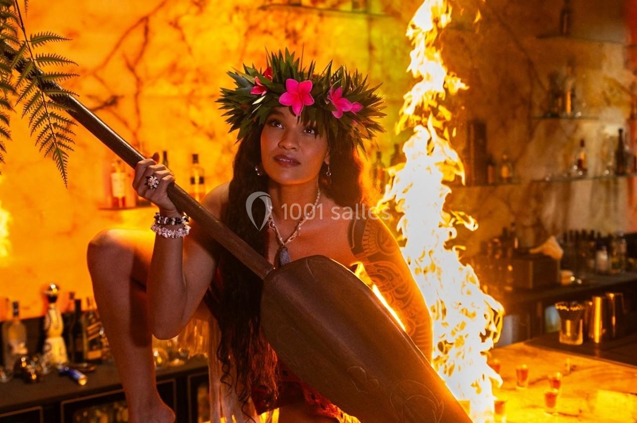 Femme en tenue traditionnelle polynésienne tenant une pagaie, devant un bar illuminé avec des flammes en arrière-plan.
