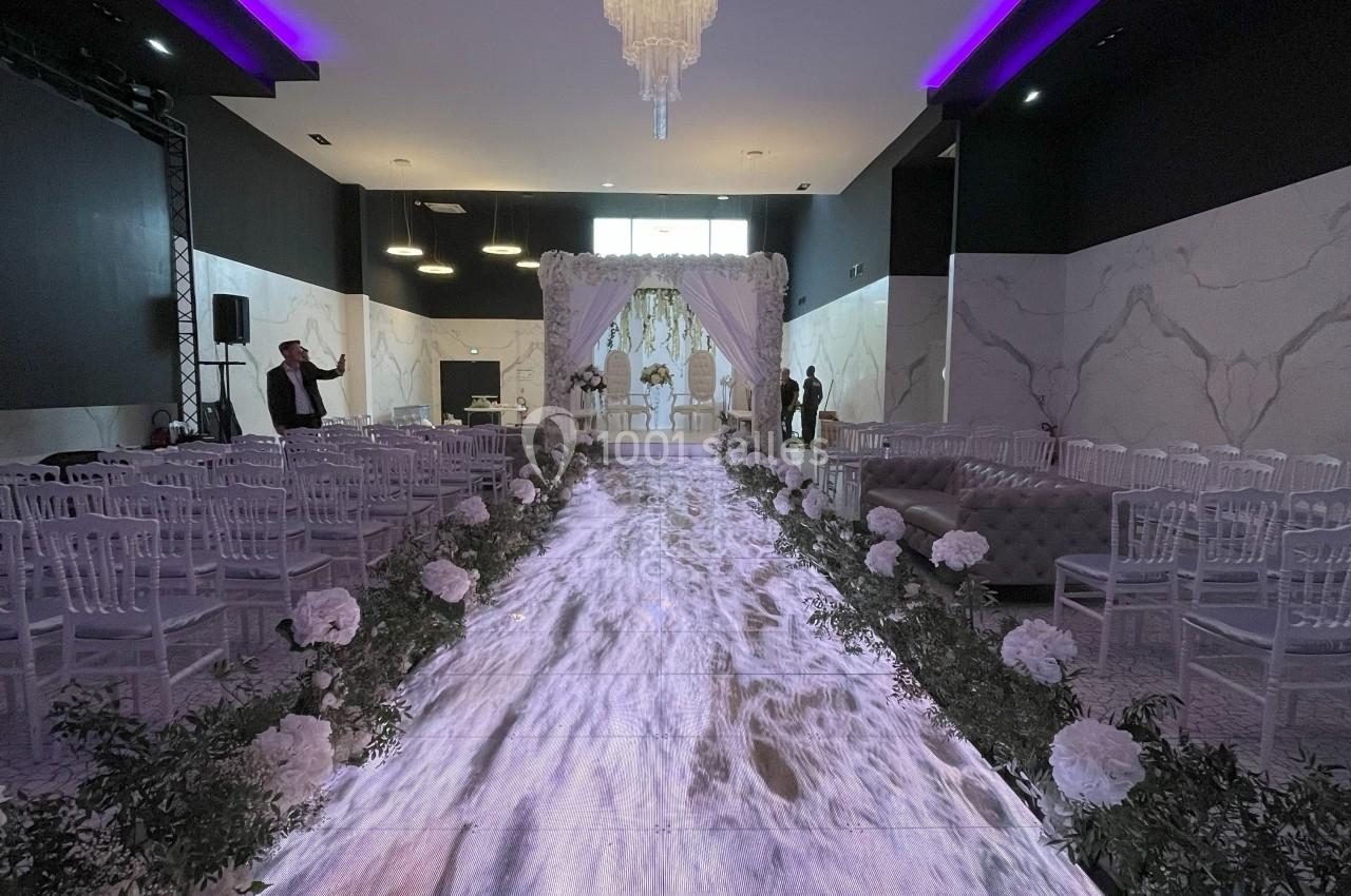 Allée centrale décorée de fleurs blanches menant à une arche nuptiale dans une salle de réception élégante.