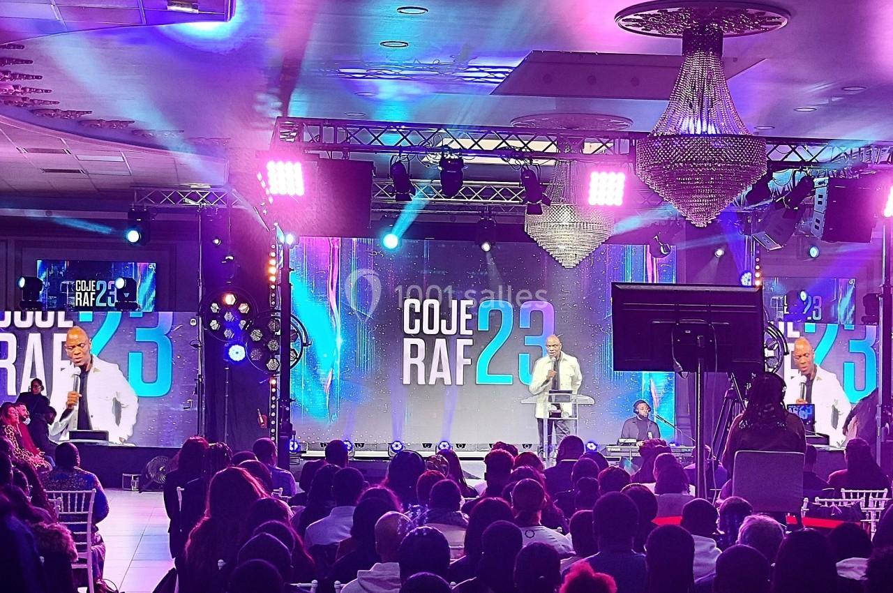 Conférence avec un orateur sur scène, éclairages colorés et public assis dans une salle équipée de projecteurs.