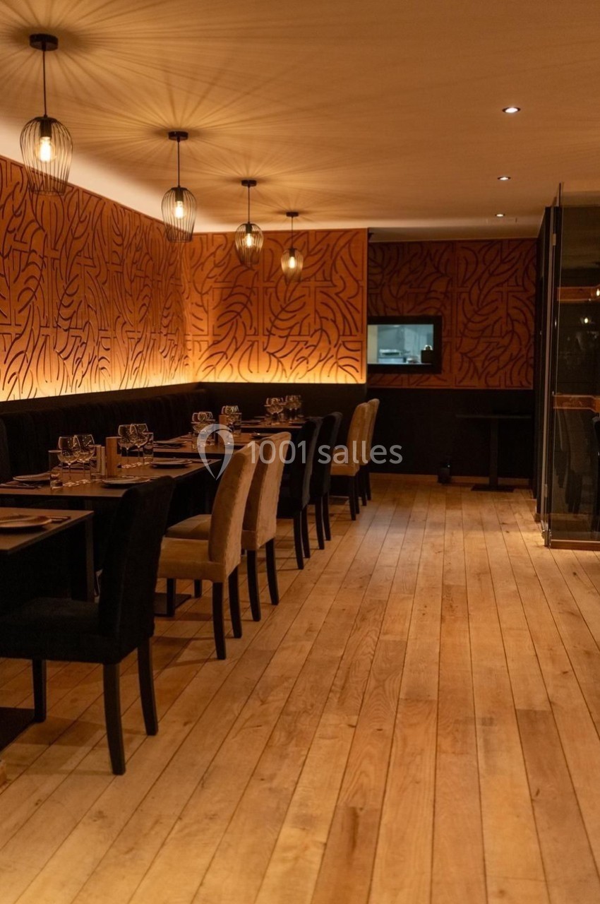 Salle de restaurant chaleureuse avec parquet en bois, murs ornés de motifs, et tables dressées sous des luminaires suspendus.